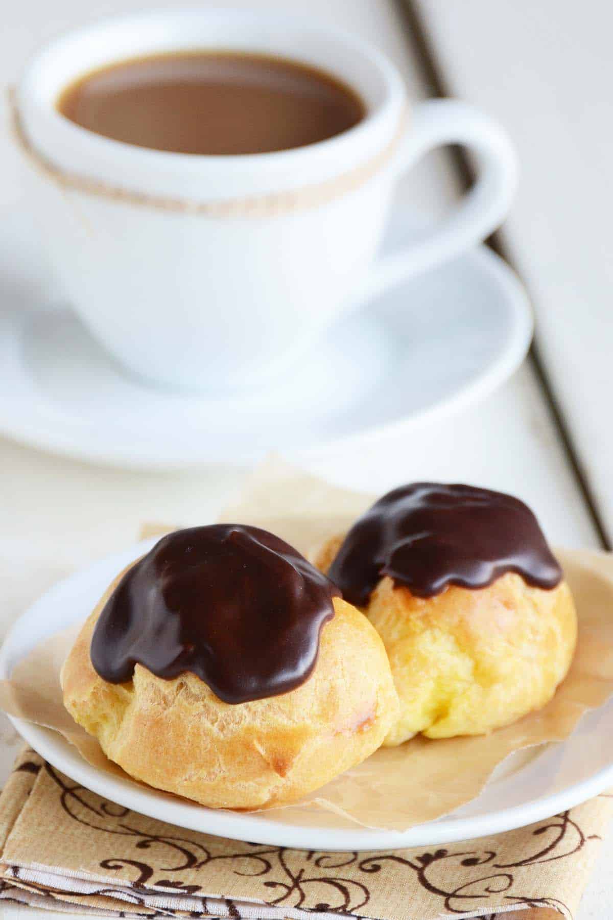 homemade cream puffs topped with chocolate on small plate with coffee in background