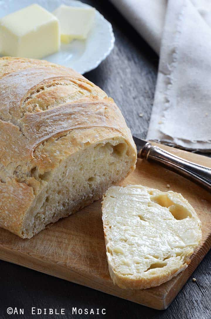 No Knead Bread Loaf on Cutting Board with a Buttered Slice of Bread