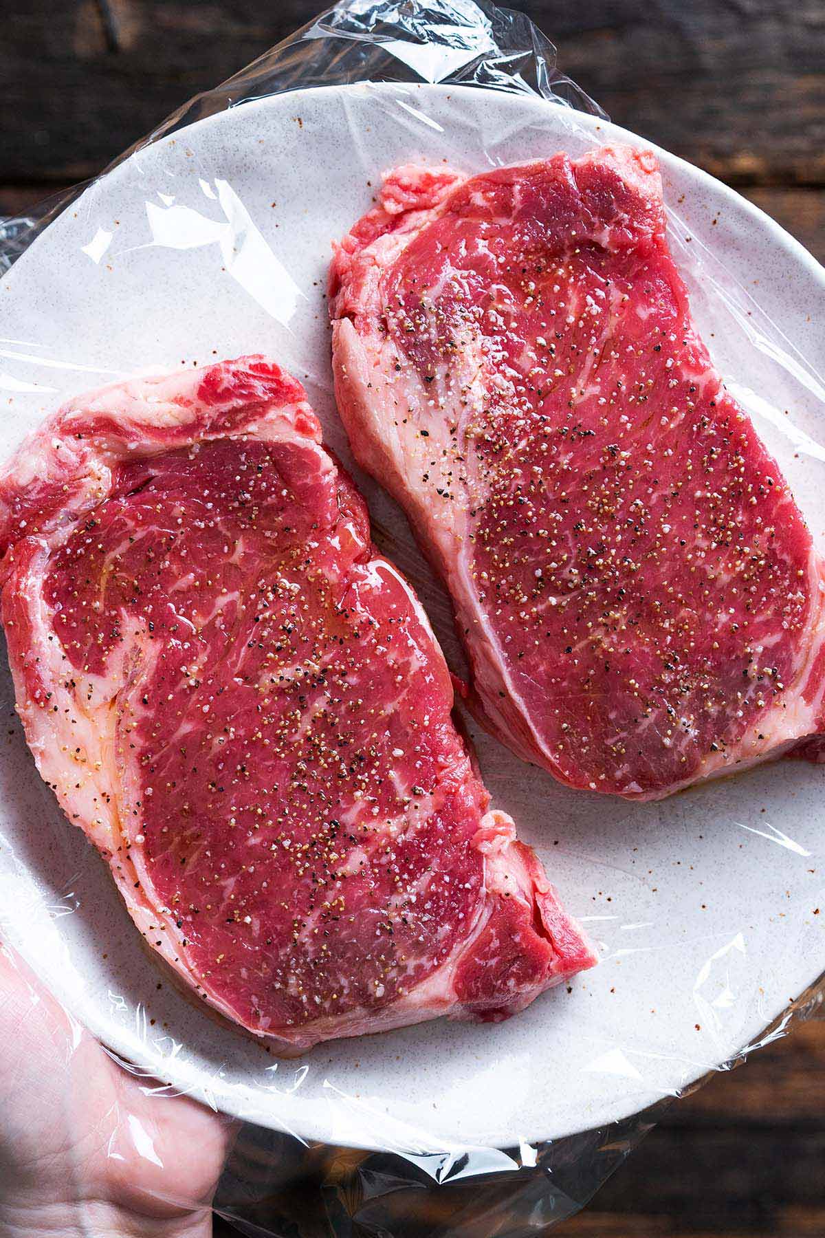 seasoned ribeyes on plastic wrap lined plate ready to grill