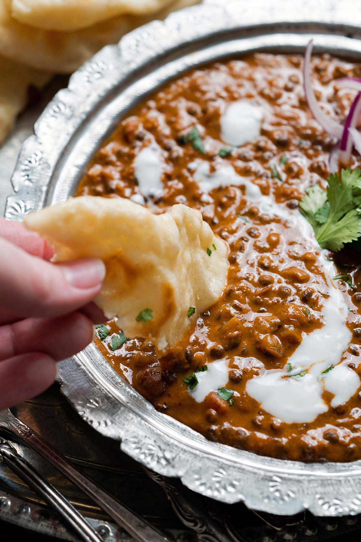 hand dipping naan bread into dish of creamy dal makhani