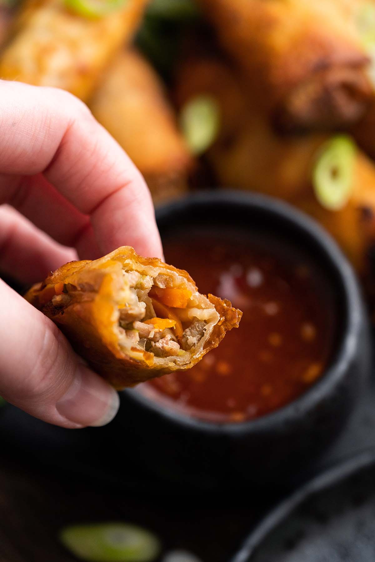 hand holding bitten egg roll over dish of sweet and sour sauce