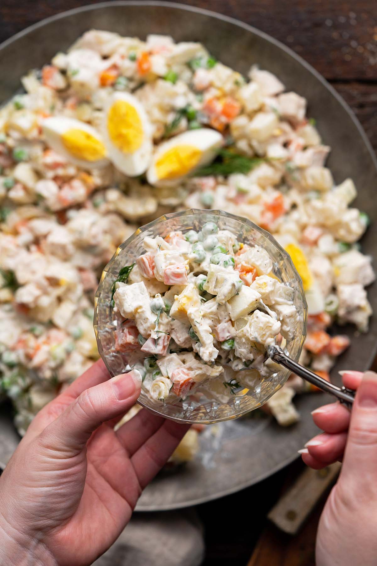 hands spooning olivier salad into glass bowl