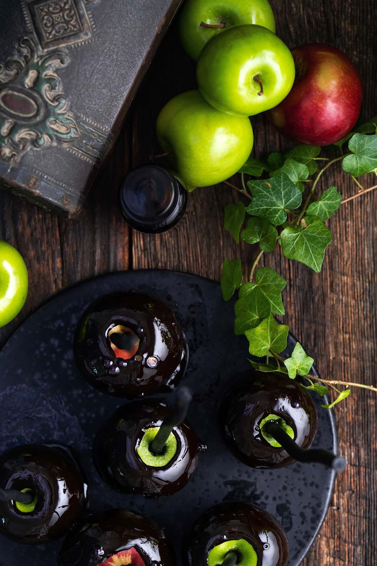 poison apples on black platter on rustic dark wooden table with ivy vine plant on side