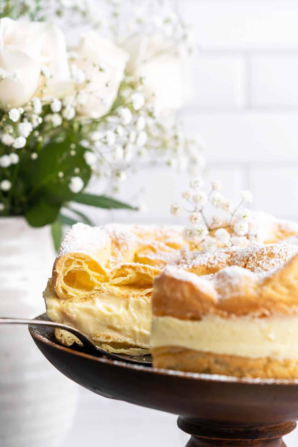 karpatka cake on wooden cake stand with boquet of white roses in background