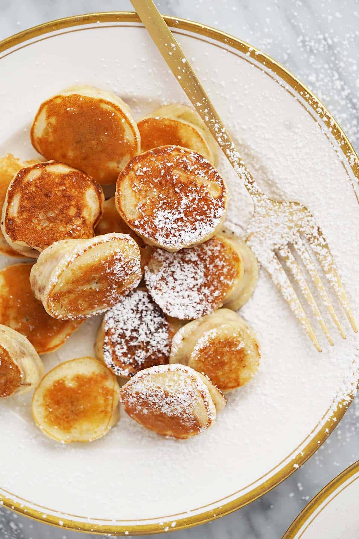 gold rimmed white plate of mini banana pancakes with powdered sugar on top