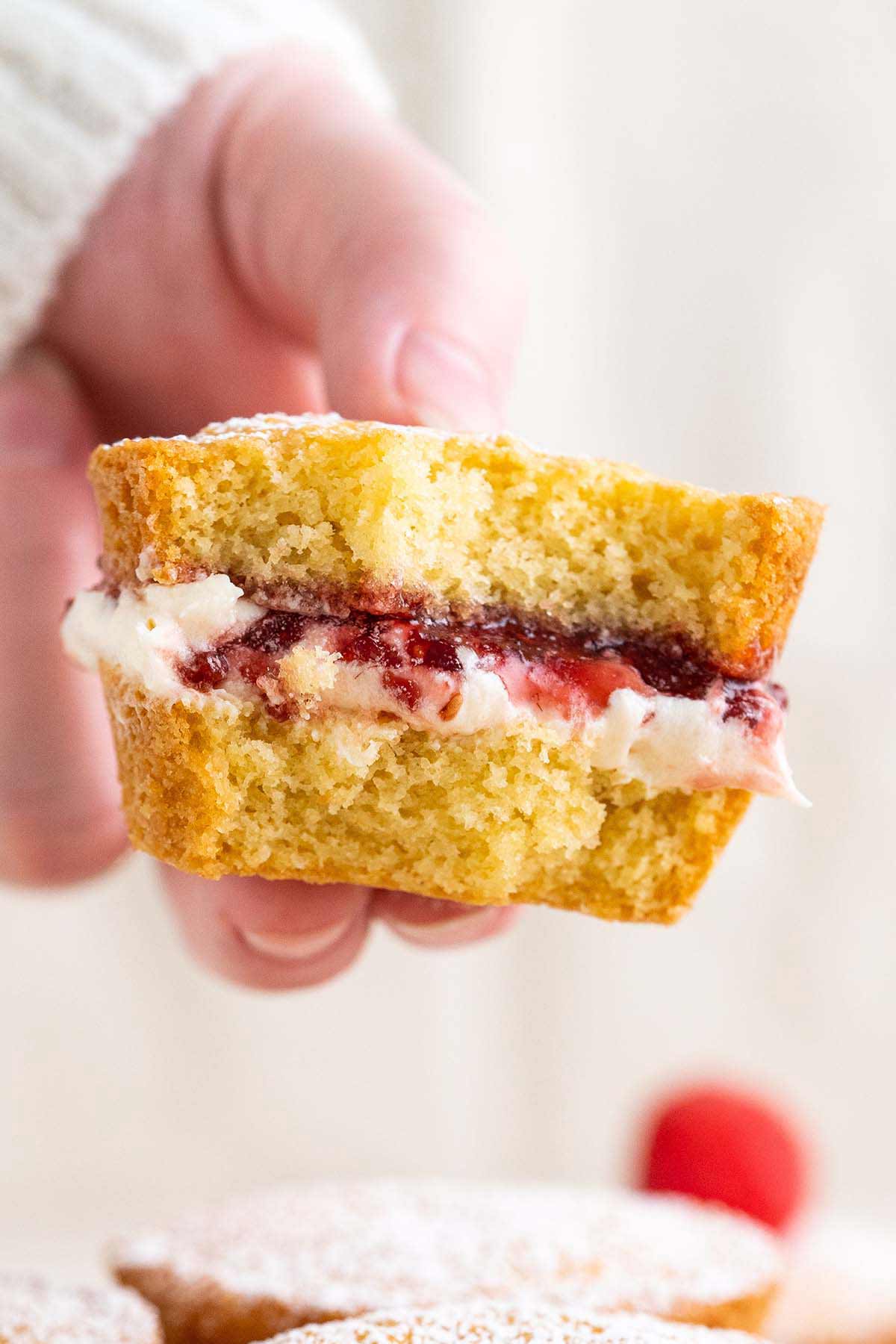 front view of hand holding half eaten mini Victoria sponge cake to show cream and jam layer in the middle