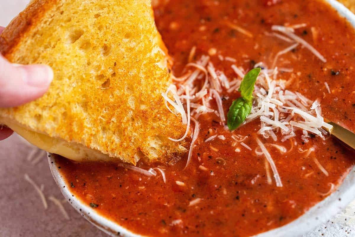 hand dipping gooey grilled cheese sandwich into homemade creamy tomato bisque