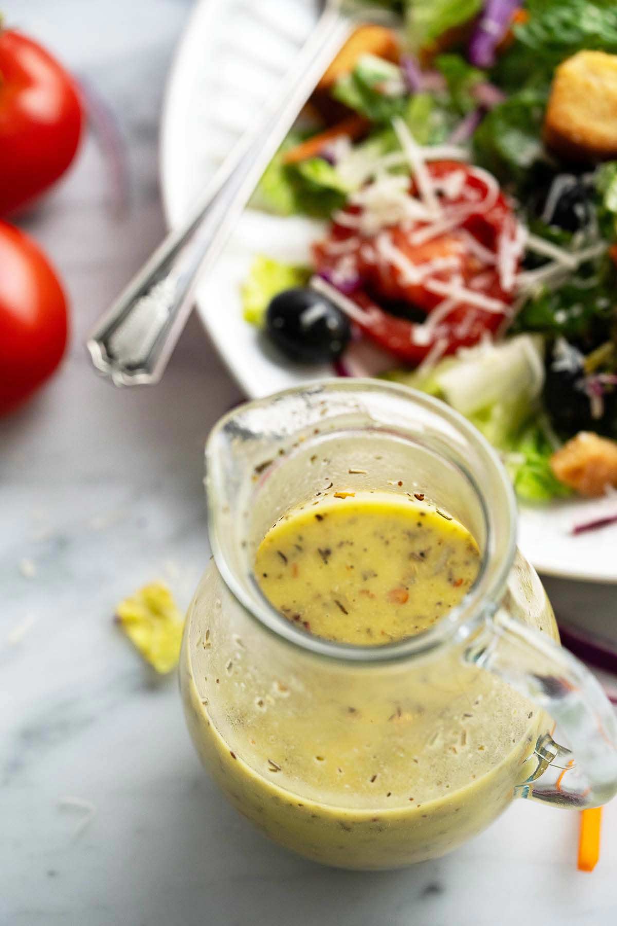 glass jug of homemade vinaigrette with salad in background on marble countertop