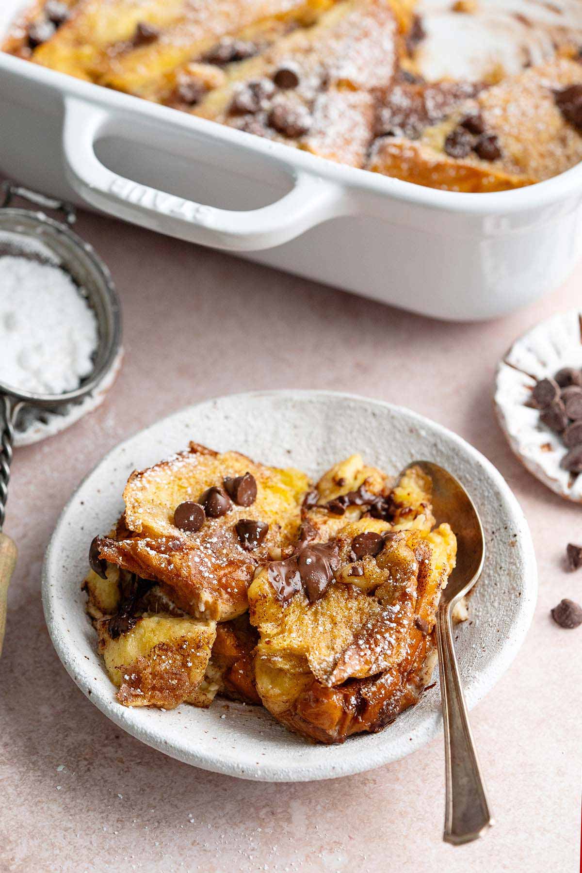 buttered bread pudding in stoneware bowl with chocolate chips and white stub casserole dish in background