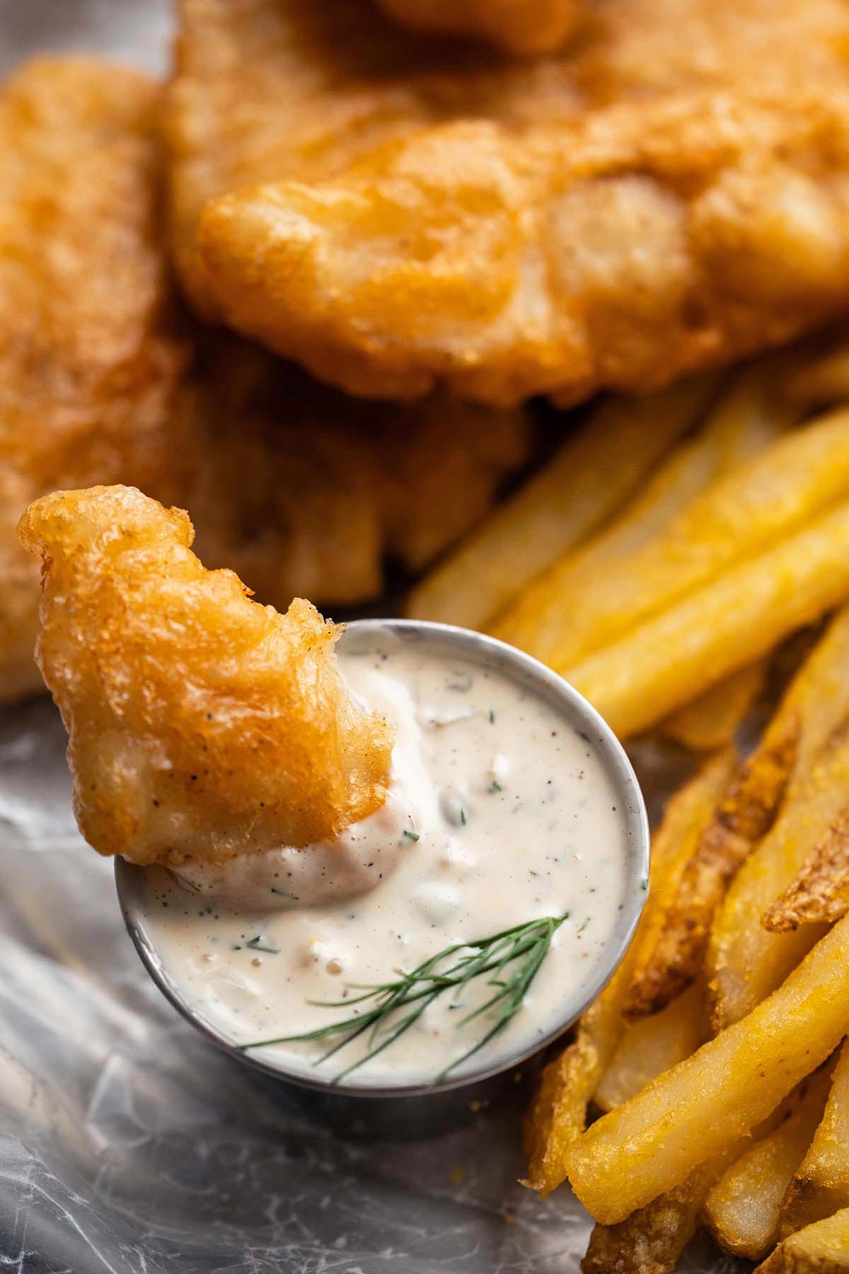beer battered fried fish dipping in small cup of creamy tartar sauce