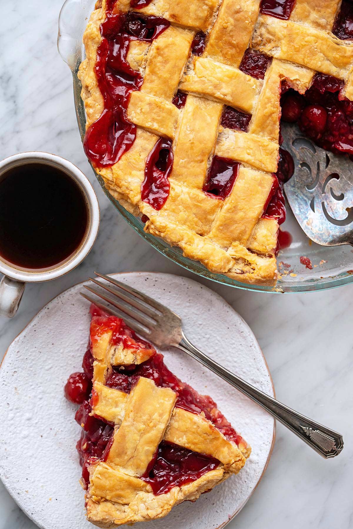slice of sour cherry pie on plate next to dish of pie and cup of coffee