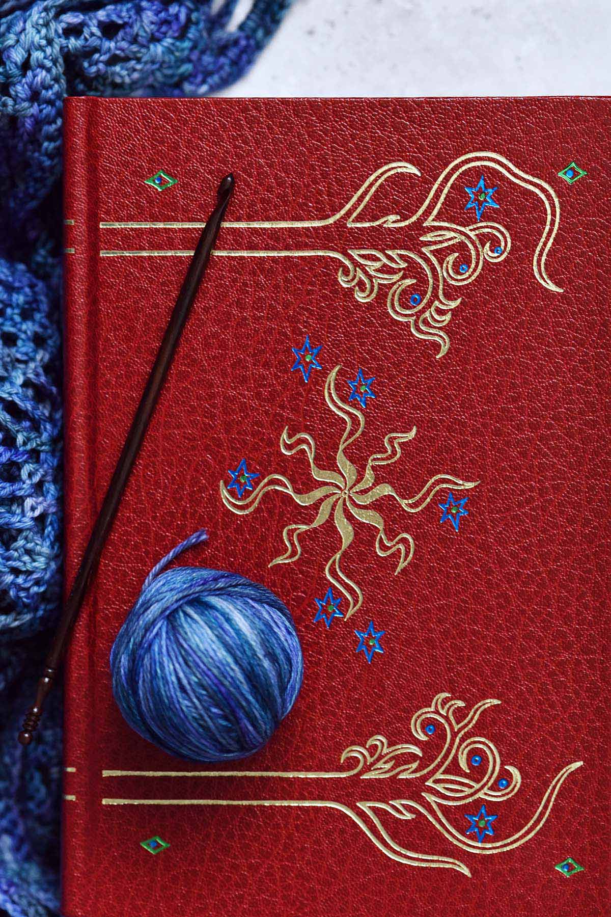 close up top view of red leather lord of the rings book with blue yarn shawl and crochet hook