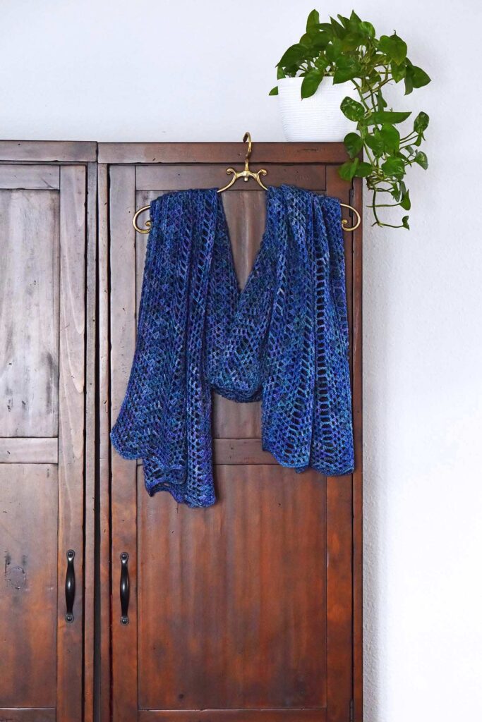 crochet rectangle shawl on brass hanger in front of wooden wardrobe with plant