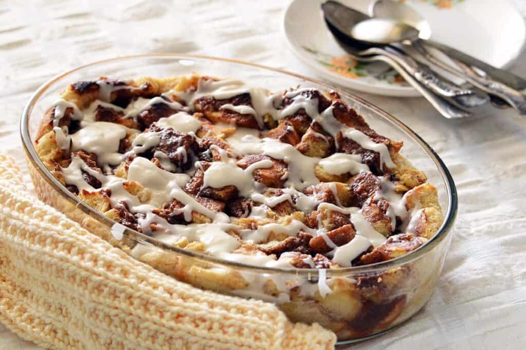 A glass dish filled with cinnamon roll bread pudding drizzled with white icing sits on a textured tablecloth. Nearby, a stack of plates with spoons and forks is visible.