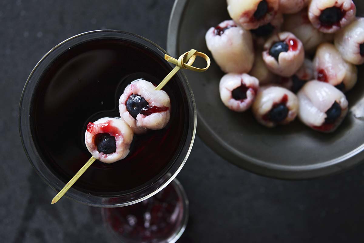 A spooky cocktail garnished with two lychee "eyeballs" skewered on a toothpick, each containing a blueberry and red syrup to resemble blood. Next to it is a bowl filled with more lychee "eyeballs.