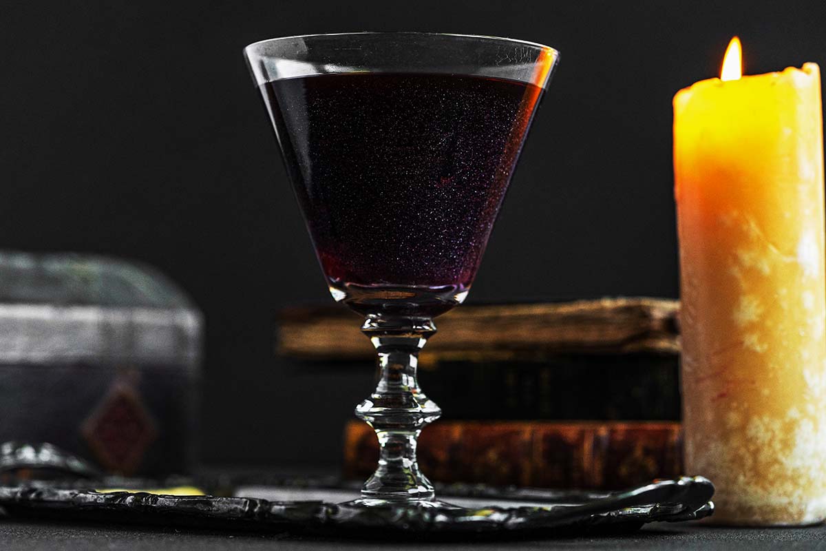 A dark, translucent drink in a stemmed glass sits on a black tray next to a lit candle. In the background, there are blurred, aged books, creating a mysterious and vintage ambiance.