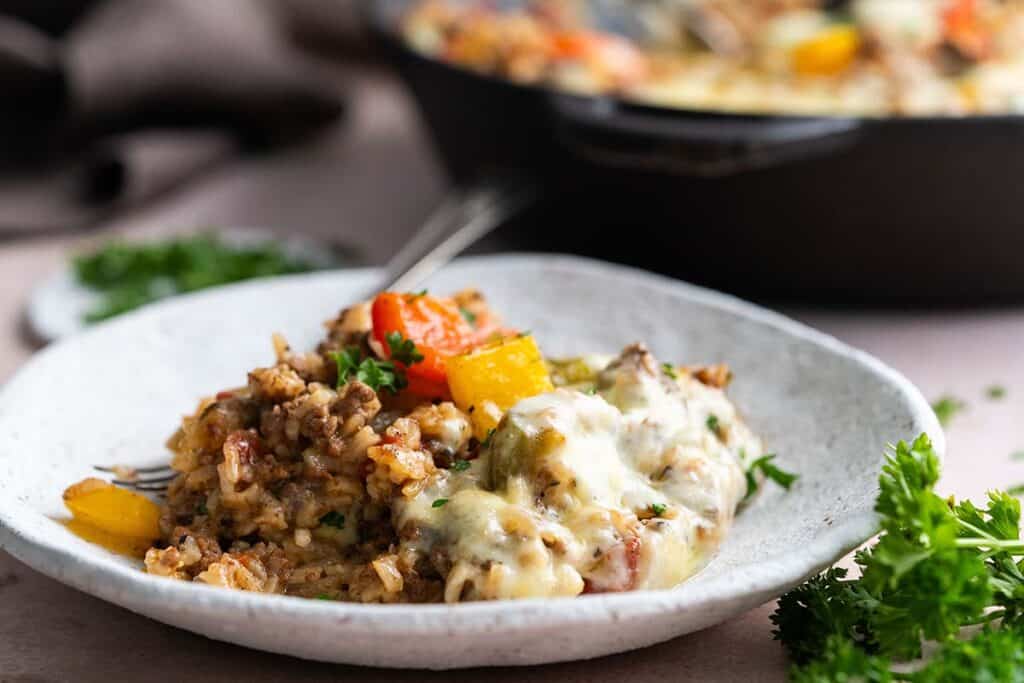 A close-up of a plate with cheesy, baked rice mixed with ground meat and colorful bell peppers. The dish is garnished with herbs, and a fork is placed beside it. A skillet with more of the dish is blurred in the background.