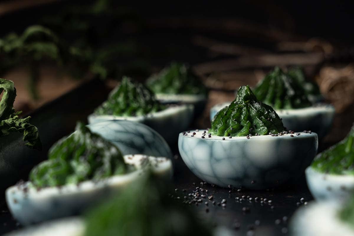 A close-up of deviled eggs with striking marbled blue and white egg whites, filled with a vibrant green herb mixture and garnished with black sesame seeds. The background is softly blurred, highlighting the intricate textures and colors of the appetizer.