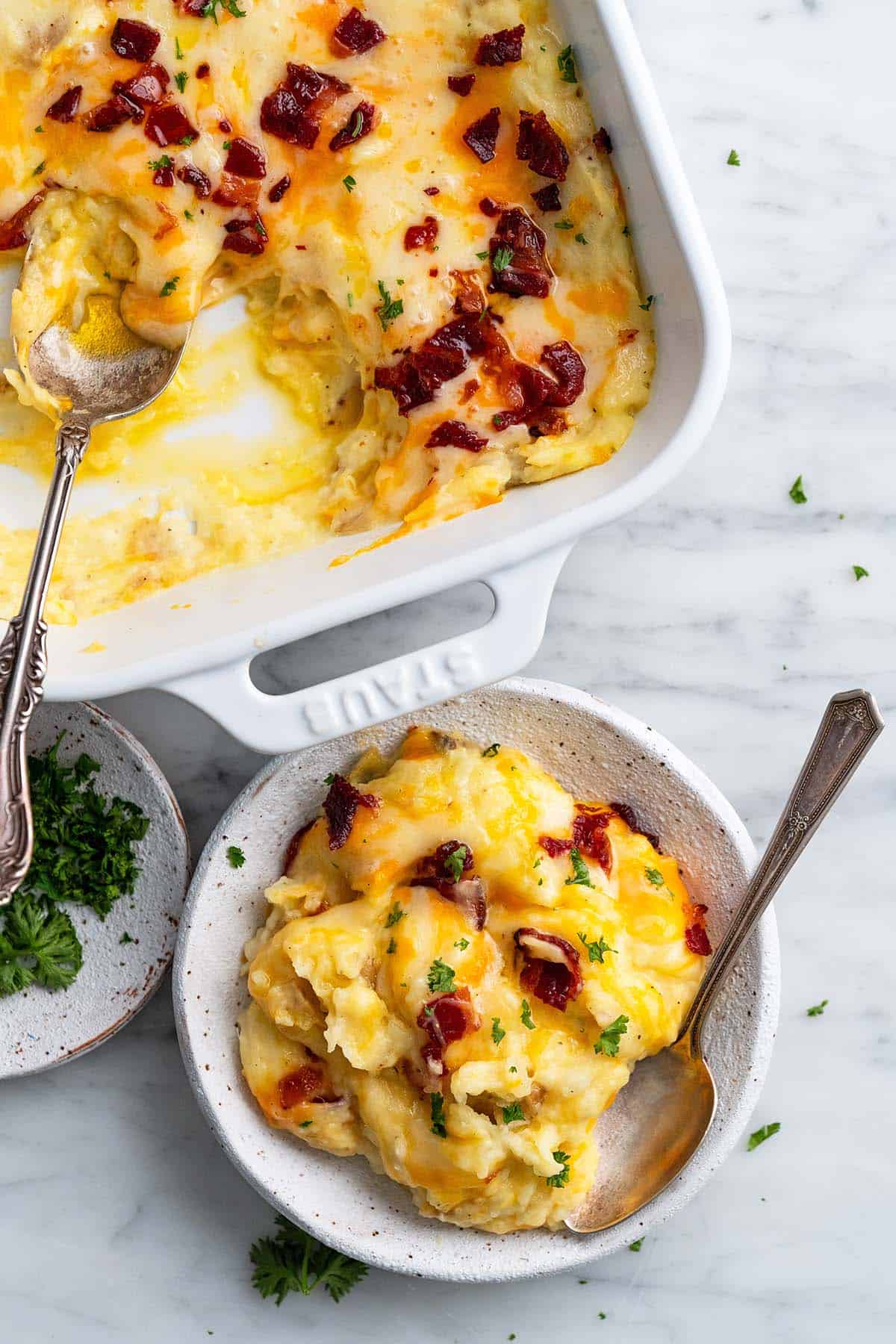 Cheesy mashed potato casserole with bacon bits and parsley garnish, served in white baking dish and individual bowls.