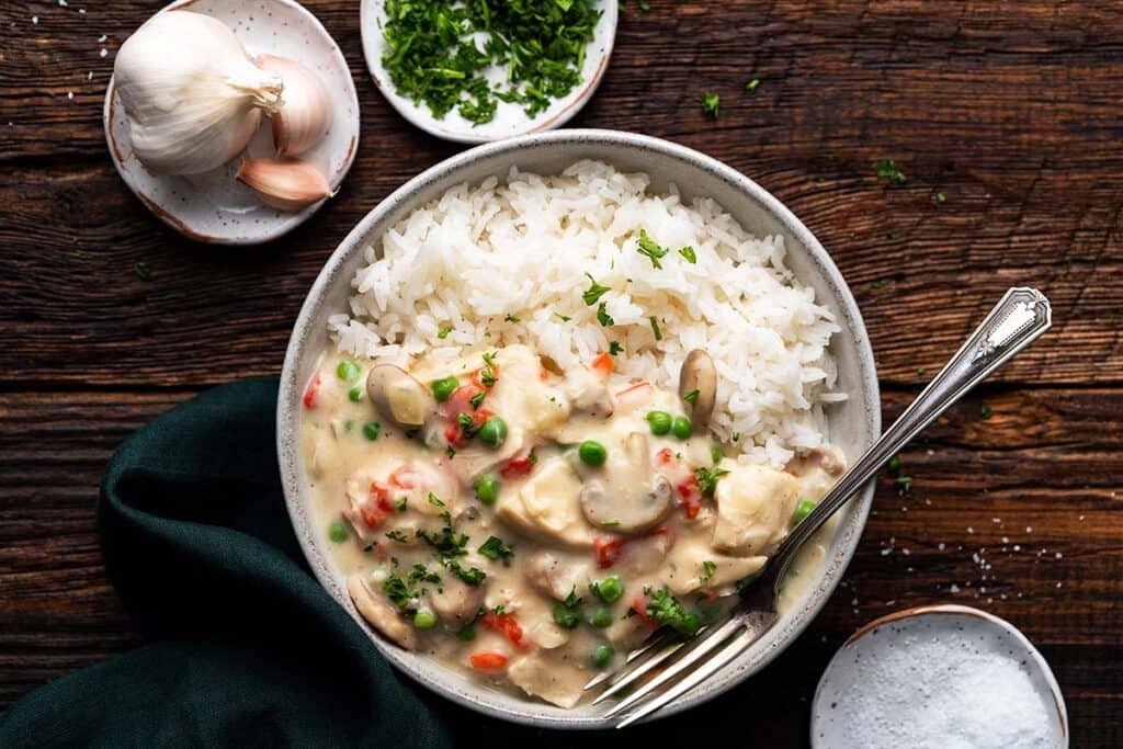 A bowl of creamy chicken à la king with sliced mushrooms, peas, and red peppers, served over white rice. Garnished with parsley. A fork rests inside the bowl. Garlic bulbs and a small dish of herbs are on the wooden table nearby.