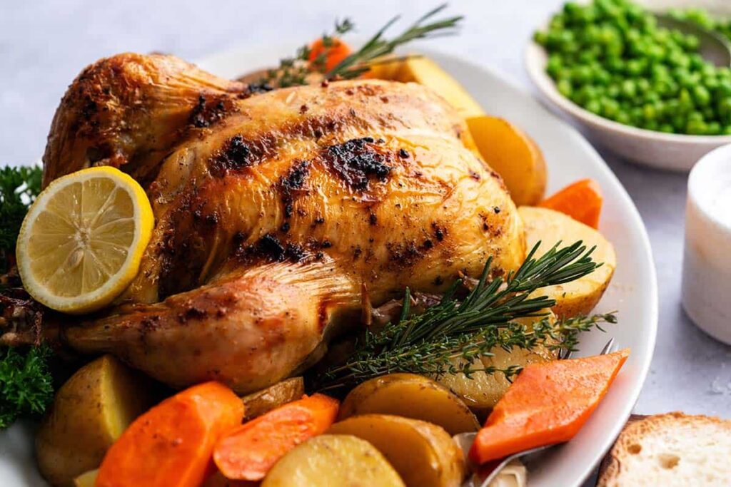 A roasted chicken garnished with herbs and a lemon slice, surrounded by roasted potatoes and carrots on a white platter. In the background, there's a bowl of green peas.