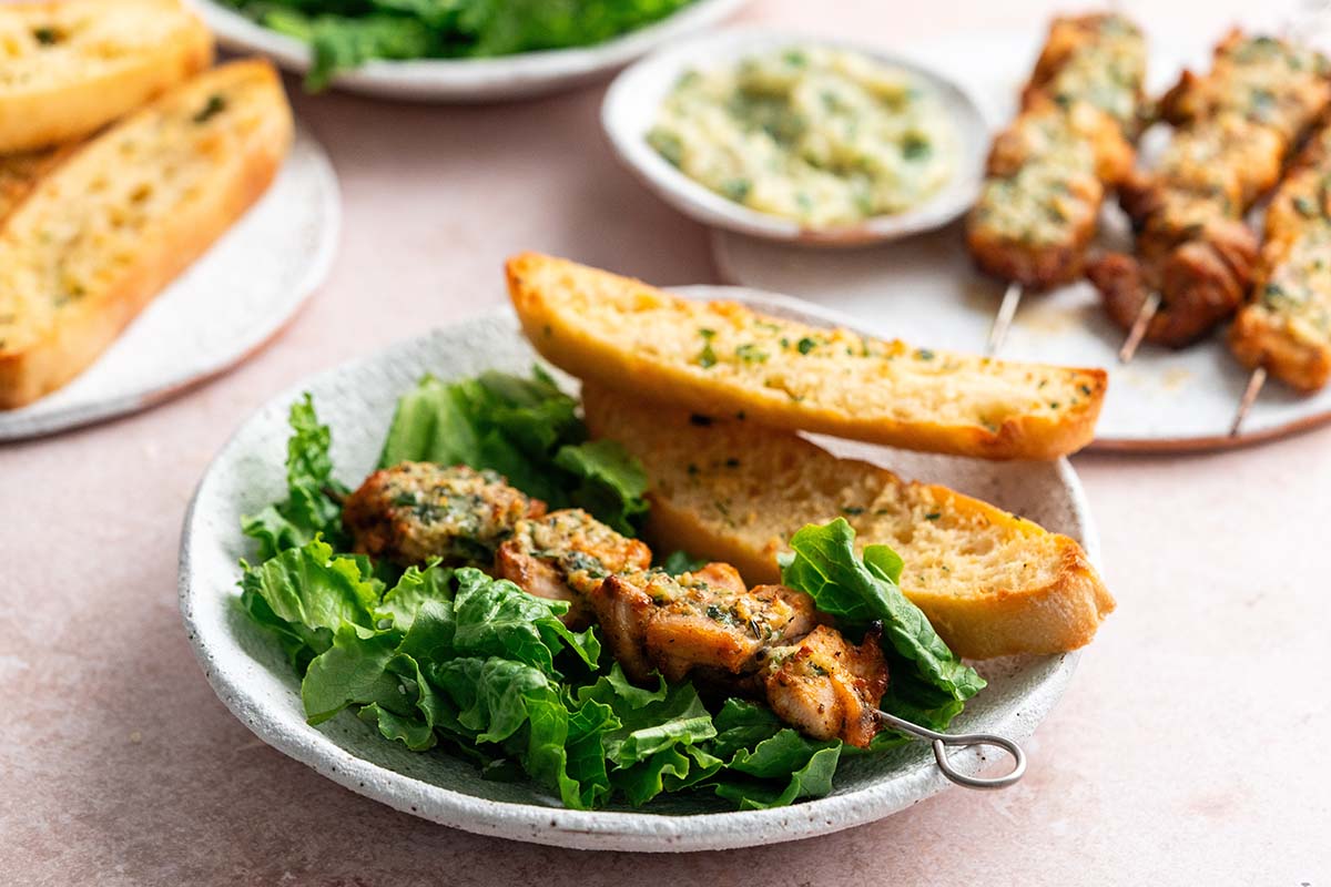 A plate with grilled chicken skewers laid on fresh lettuce, accompanied by two pieces of garlic bread. In the background, there are additional plates with more garlic bread, a dip, and salad greens.
